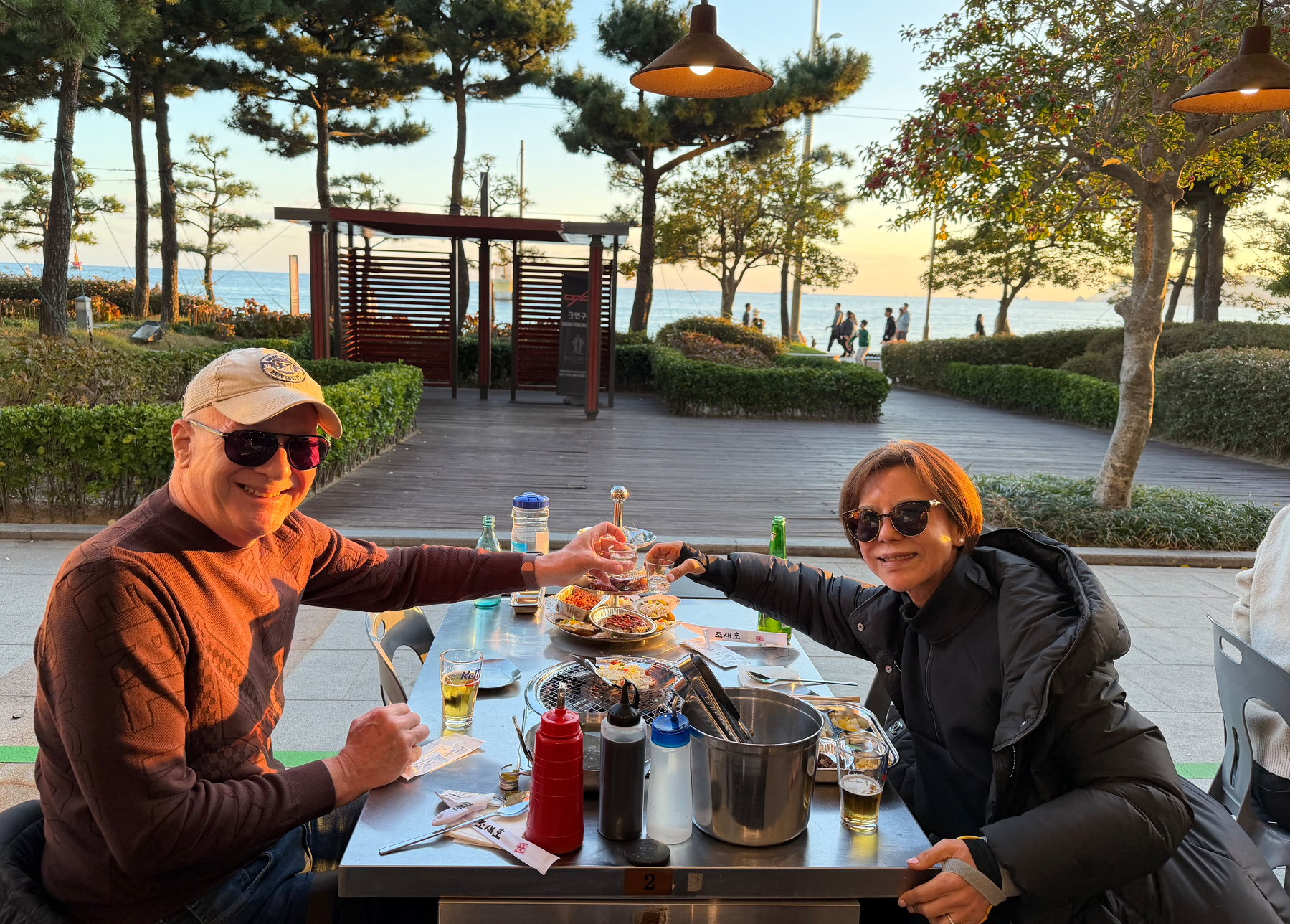 Auto-generated description: A smiling couple wearing sunglasses enjoys a meal at an outdoor restaurant near a waterfront, surrounded by trees and a sunset view.