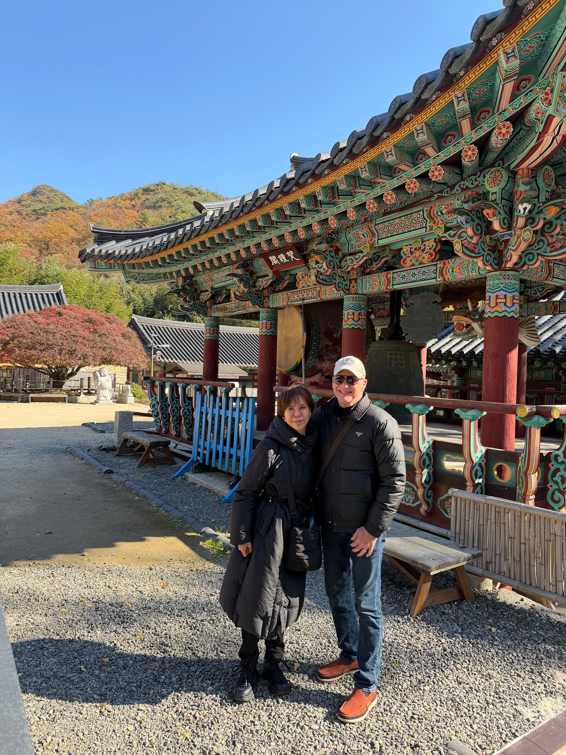 Auto-generated description: A couple is posing in front of a traditional Korean temple with colorful architecture and autumn trees in the background.